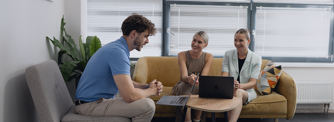 Cross-functional team having a friendly conversation in a casual office lounge with laptops on a coffee table.