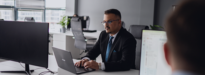 Well-dressed focused developer coding on a laptop at a shared desk in a modern office setting.