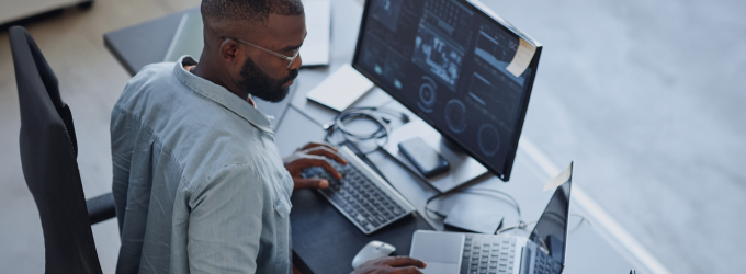 Developer analyzing data dashboards on dual monitors while writing code at the desk.