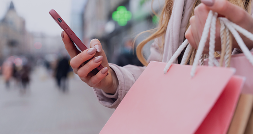 Consumer checks online deals on smartphone amid pink shopping bags and urban storefront backdrop.